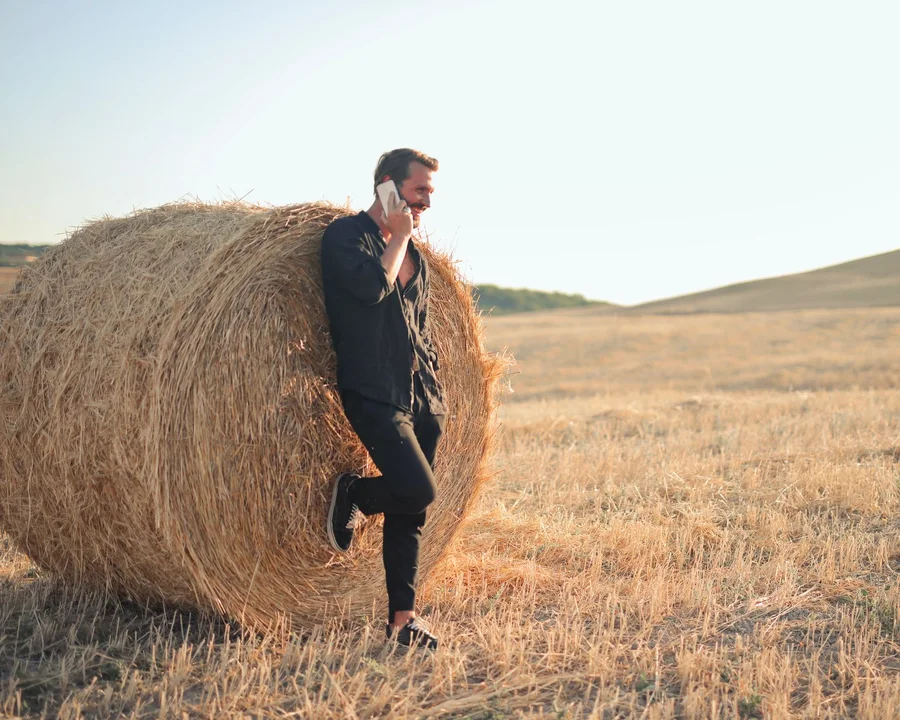 A man on his mobile, leaning on a hay bale in a dry paddock